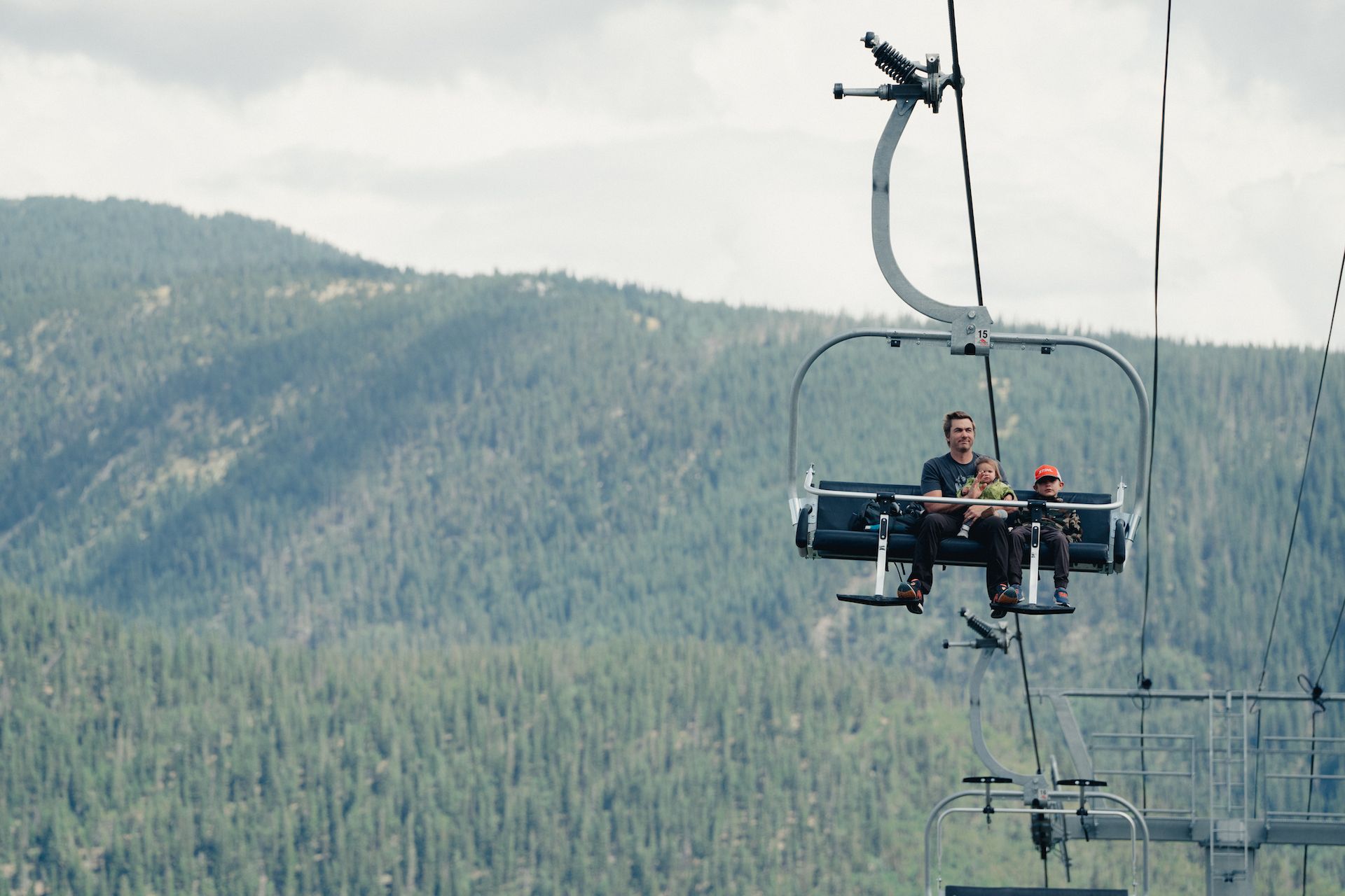 Two people riding a chairlift in the summer with Taos Ski Valley in the background.
