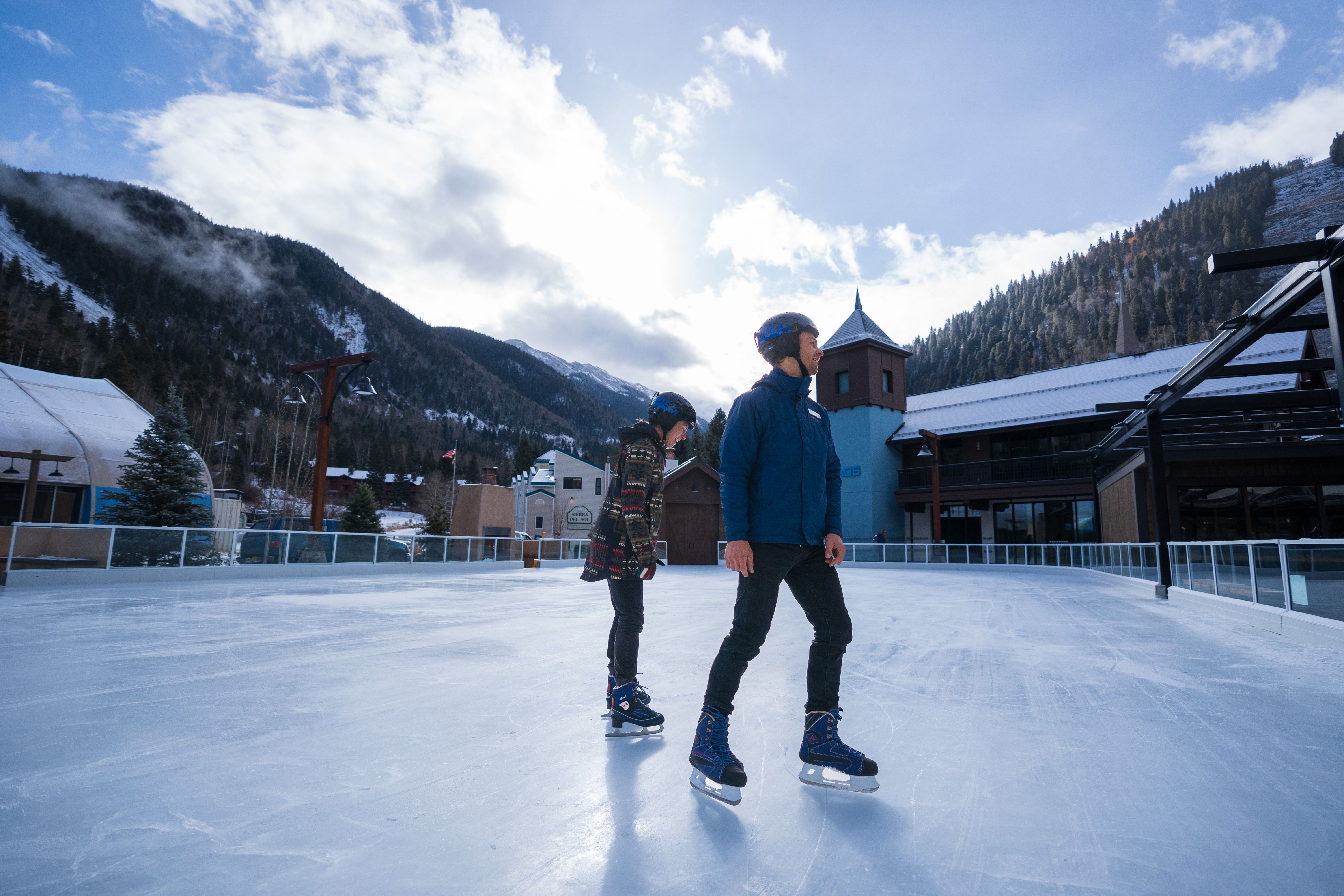 Two people ice skate on a rink in Taos Ski Valley on a sunny day.